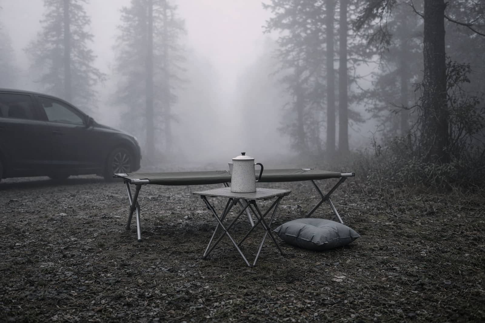 A folding cot, a roll-top table, an inflatable pillow and an enamel percolator at a misty car campsite