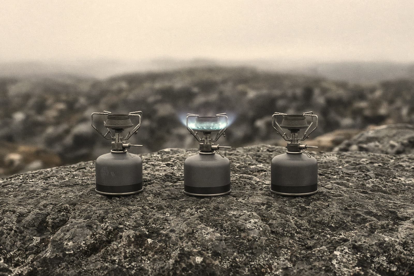 Three small canister stoves lined up on a flat rock, one lit with a low blue flame