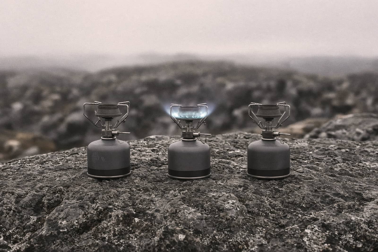 Three small canister stoves lined up on a flat rock, one lit with a low blue flame
