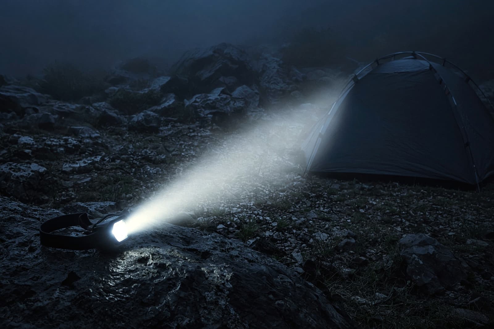 A headlamp resting on a rock throwing a beam through light mist across a dark tent site at blue hour