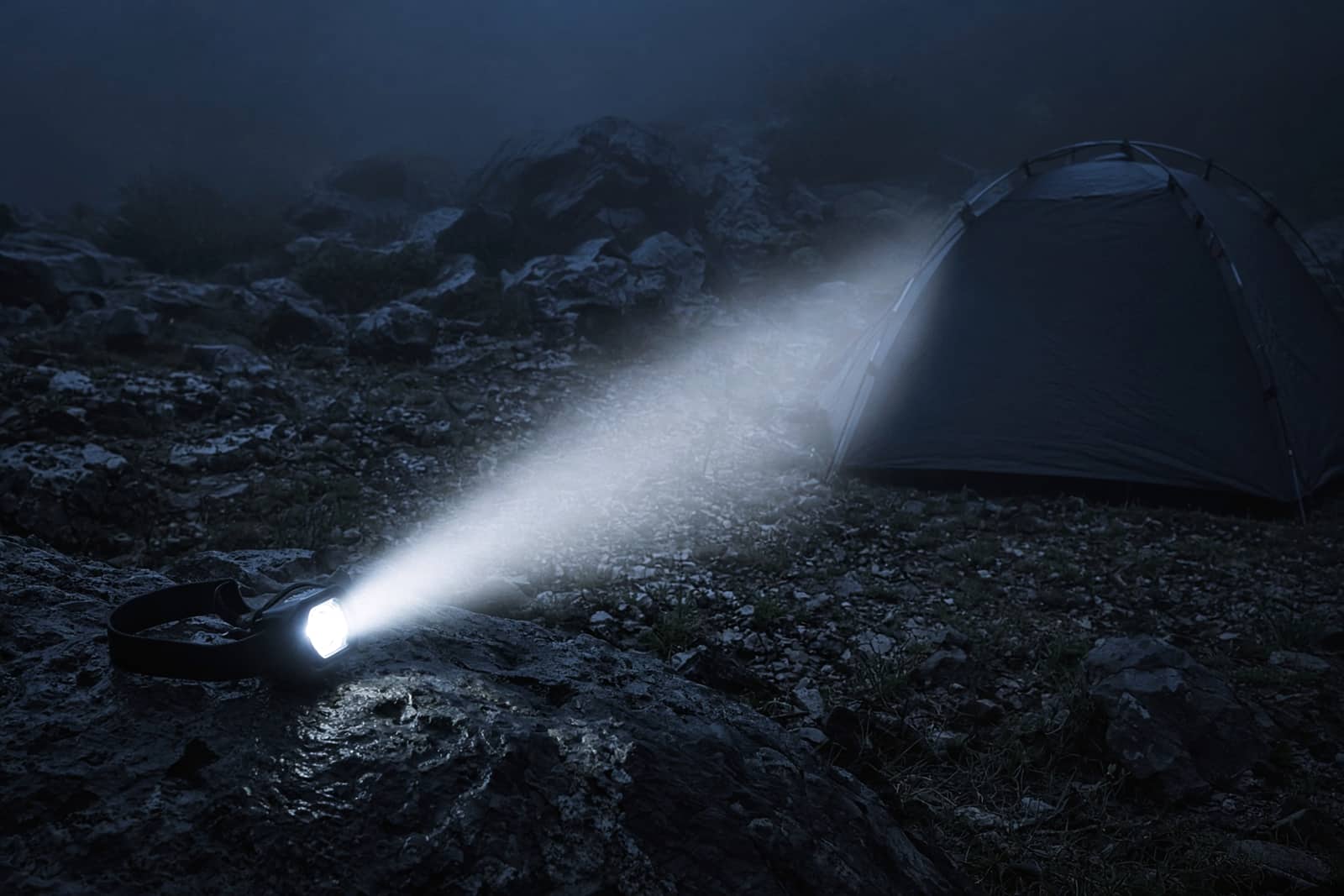 A headlamp resting on a rock throwing a beam through light mist across a dark tent site at blue hour