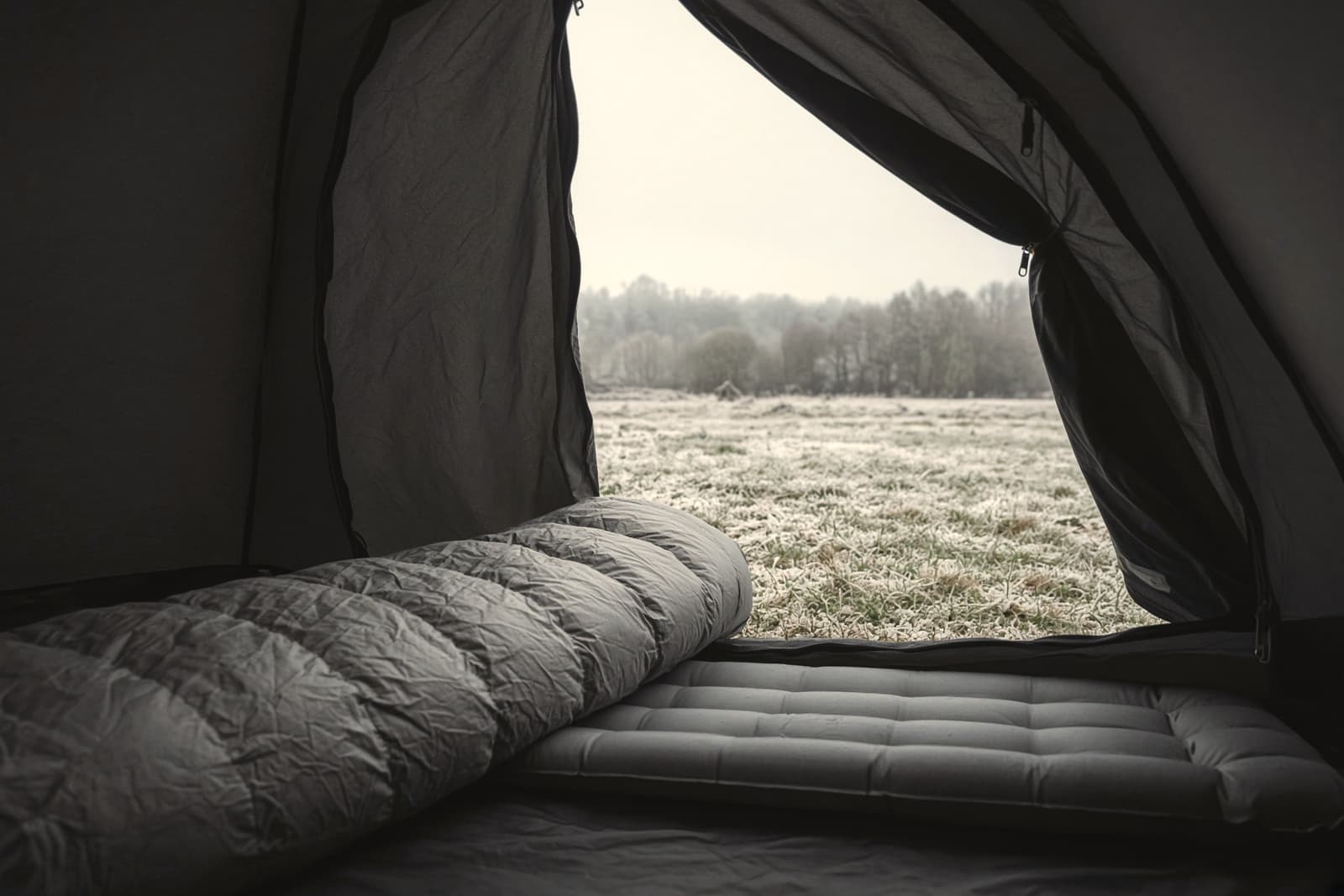 A lofted down mummy sleeping bag on a pad seen through an open tent door, frost on the grass outside, early morning