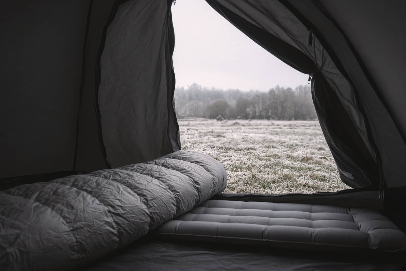 A lofted down mummy sleeping bag on a pad seen through an open tent door, frost on the grass outside, early morning