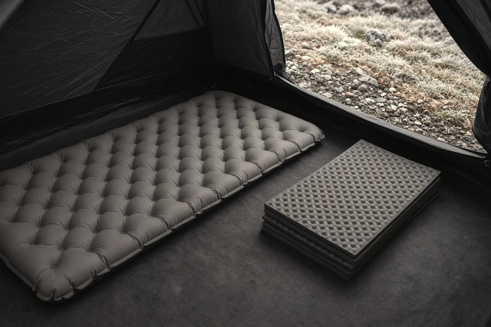An inflated air sleeping pad beside a folded closed-cell foam pad on a tent floor, frosty ground visible at the door,