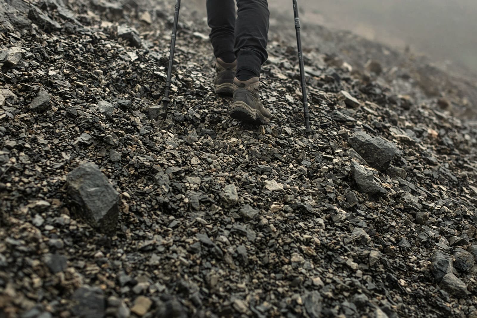 A pair of trekking poles planted on a steep rocky descent, hiker visible from the knees down, loose scree