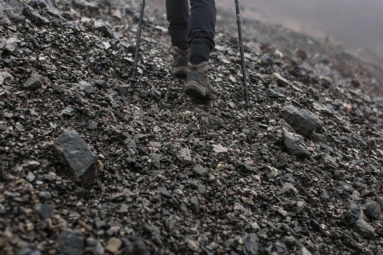 A pair of trekking poles planted on a steep rocky descent, hiker visible from the knees down, loose scree