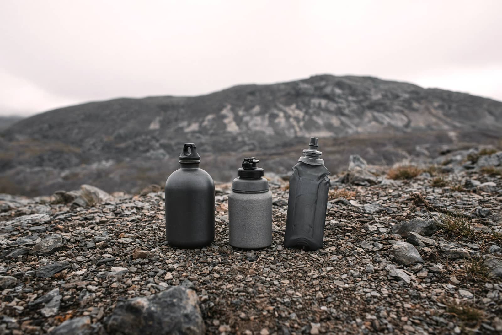 Two bottles and a soft flask on a dry rocky trail with a far ridge
